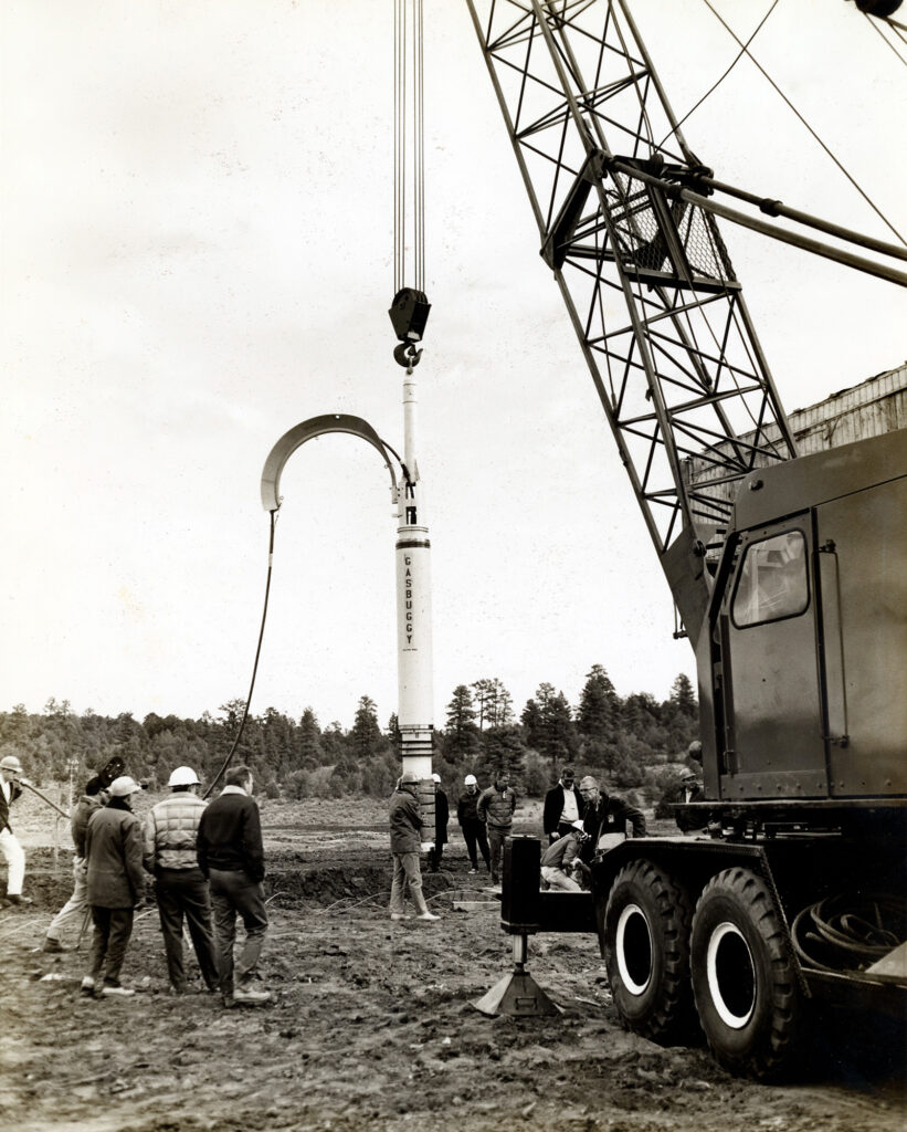 Black and white photo of men around a missile-shaped object being lowered into a hole by crane