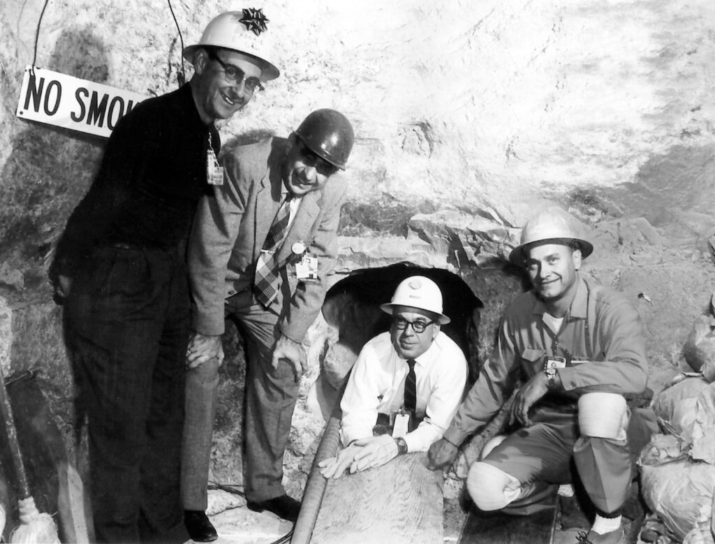 Four men in hardhats smiling and posing inside a well-lit cave, one lying on his belly halfway inside a narrow hole.