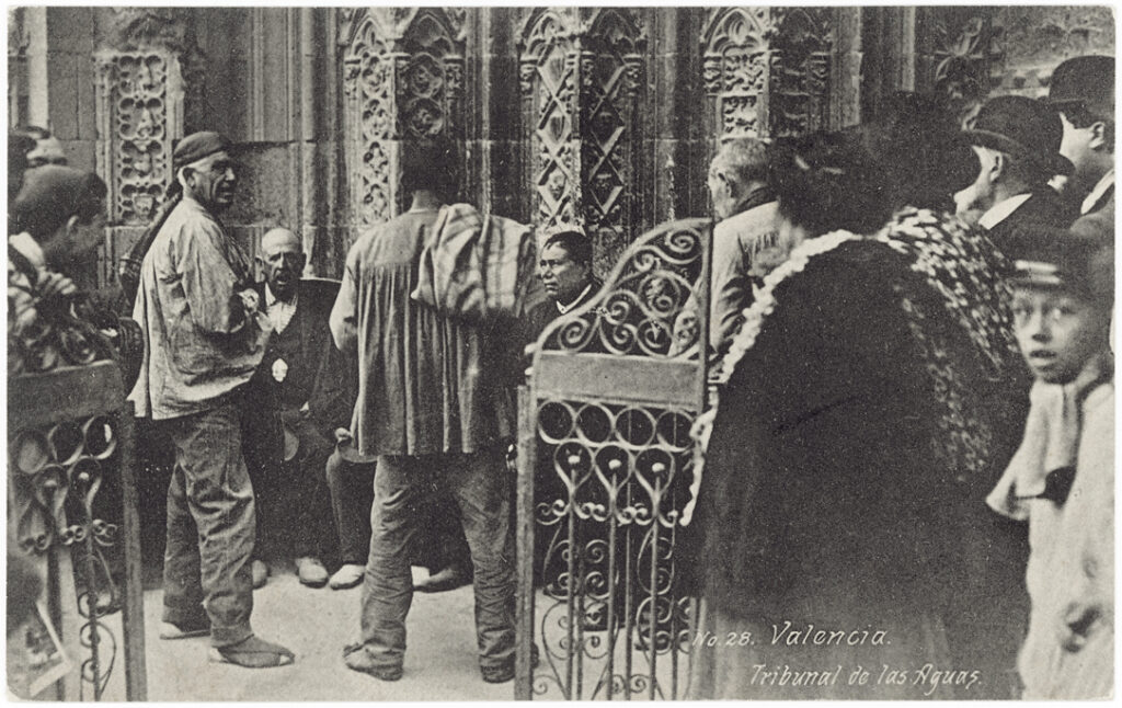 Old photo of two plain-dressed men standing before a circle of well-dressed men seated by cathedral doors while a crowd looks on from behind iron gates