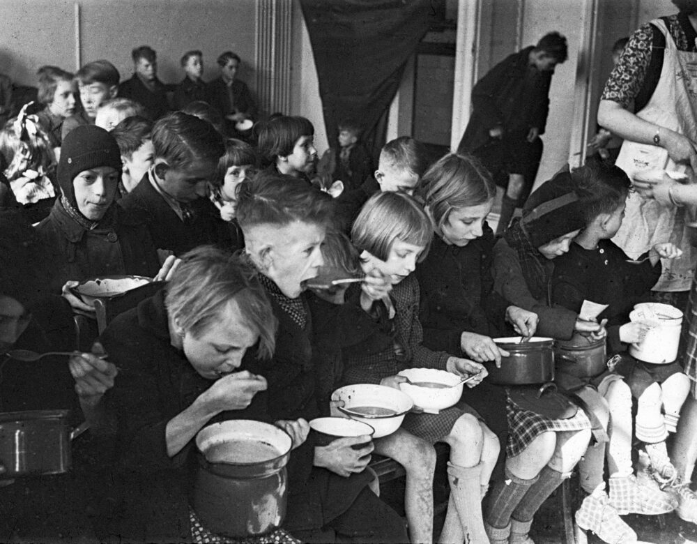 Black and white photo of dozens of children sitting with bowls of soup