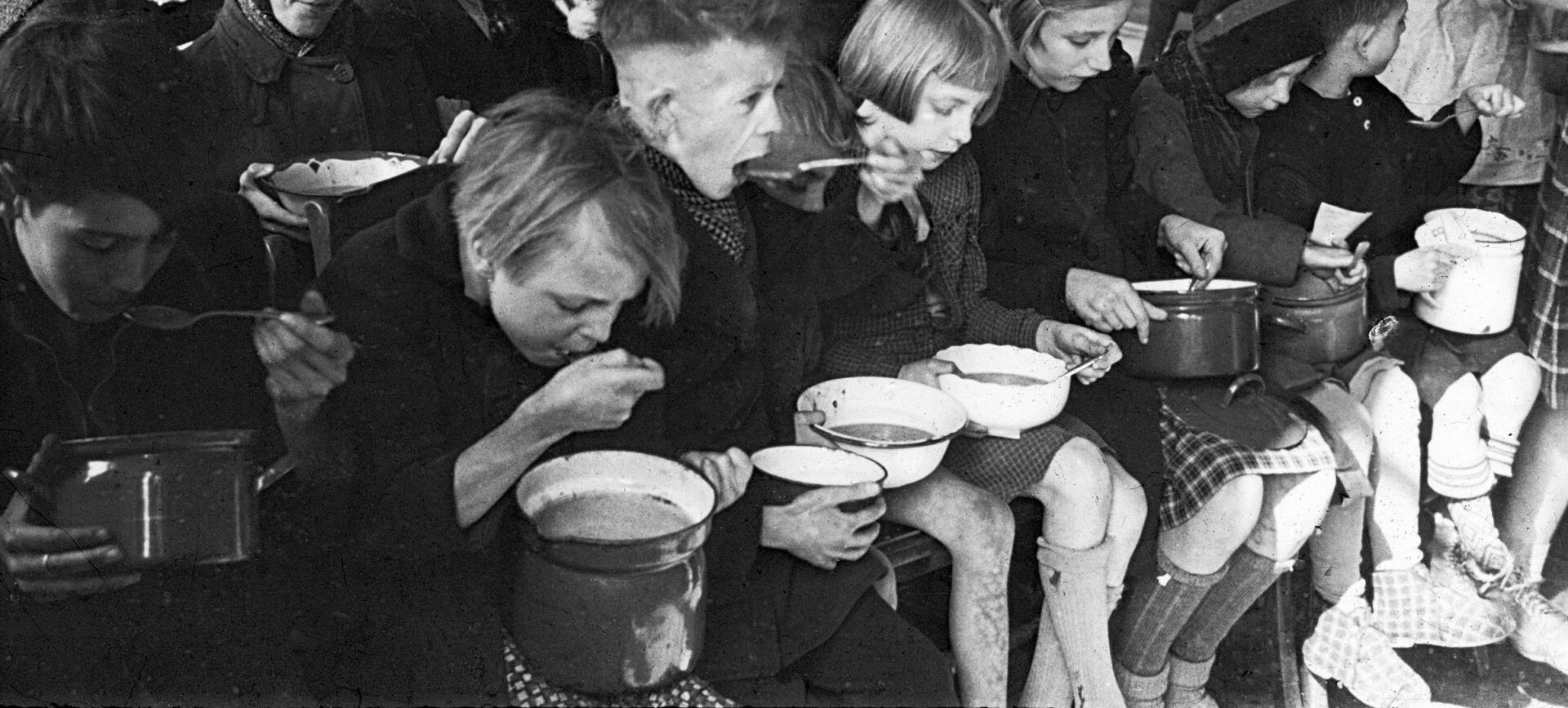 Black and white photo of dozens of children sitting with bowls of soup