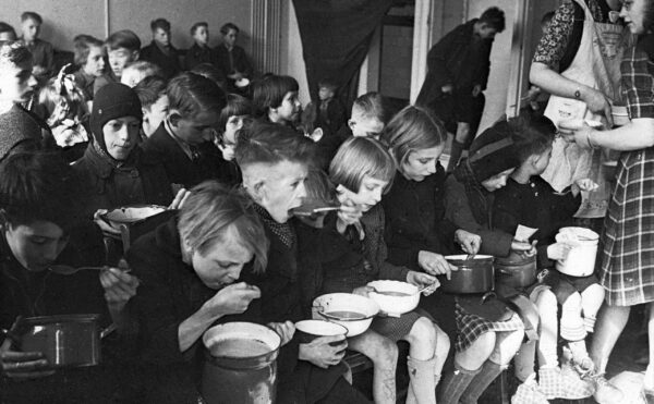 Black and white photo of dozens of children sitting with bowls of soup