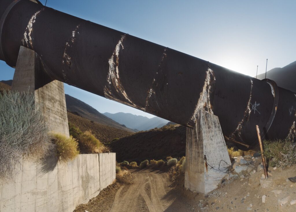 Large, rusty pipe on concrete supports with dirt road underneath