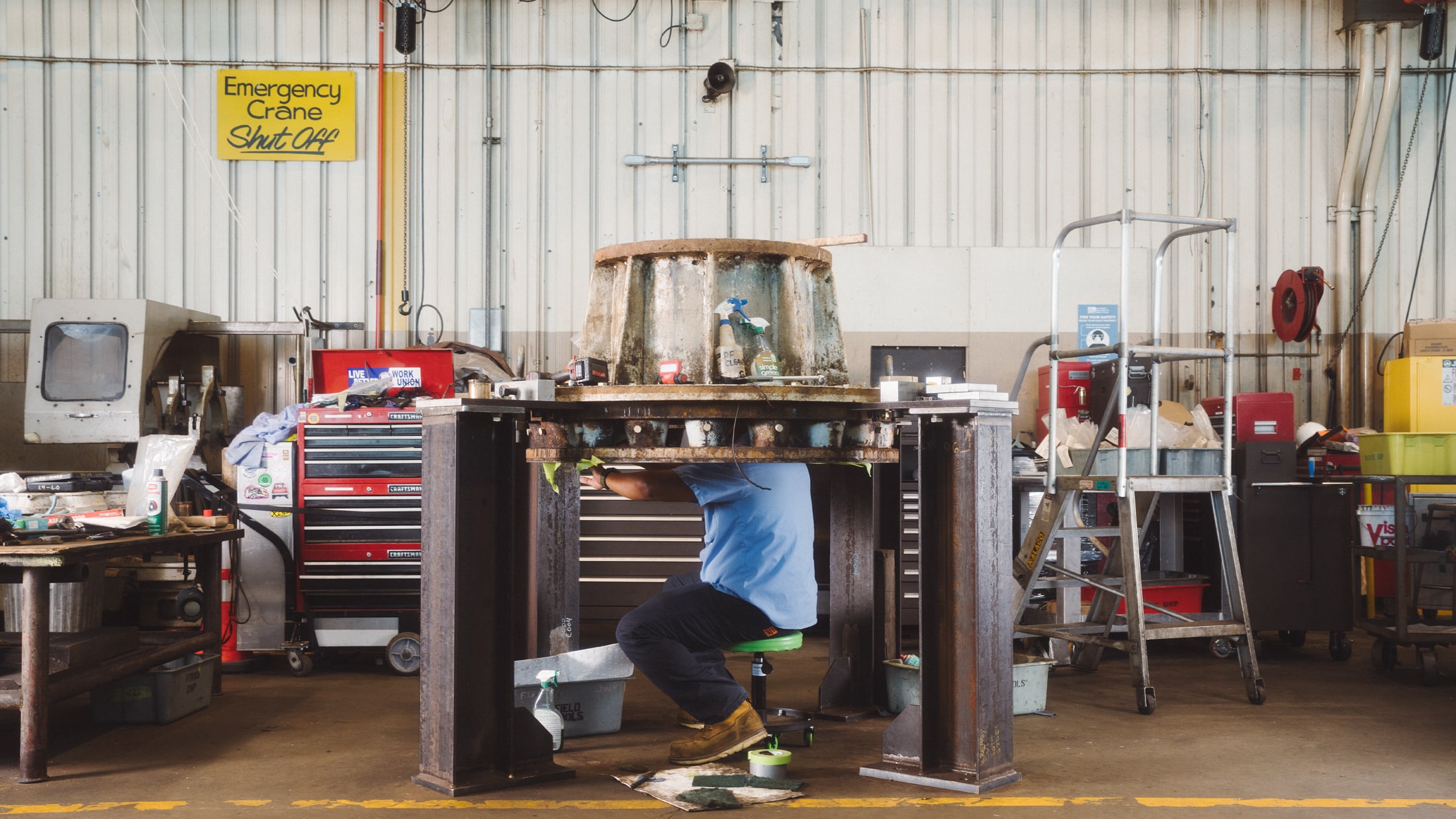 Man squatting underneath a large piece of metal equipment, his head obscured