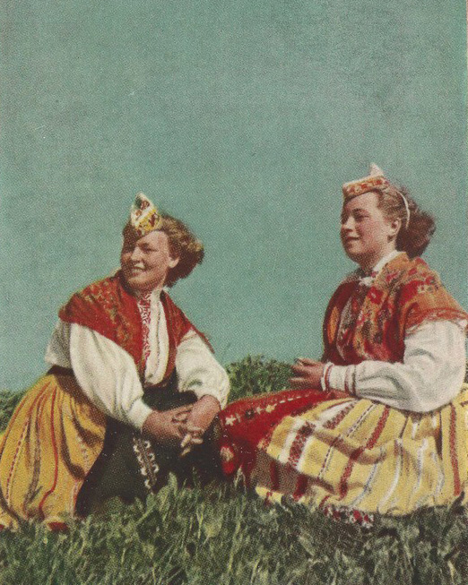 Old color photo of two women in traditional Estonian clothing sitting in grass