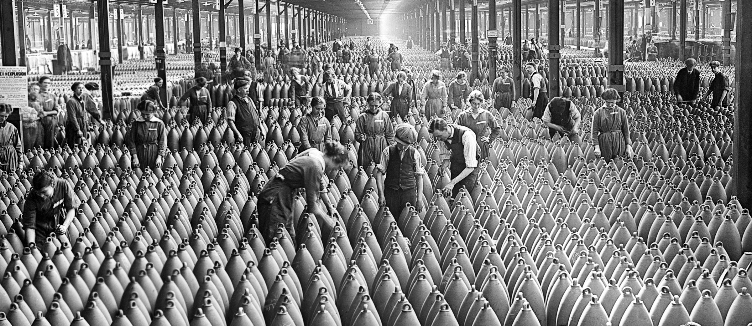 Old photo showing men and women working in a large factory warehouse full of artillery shells