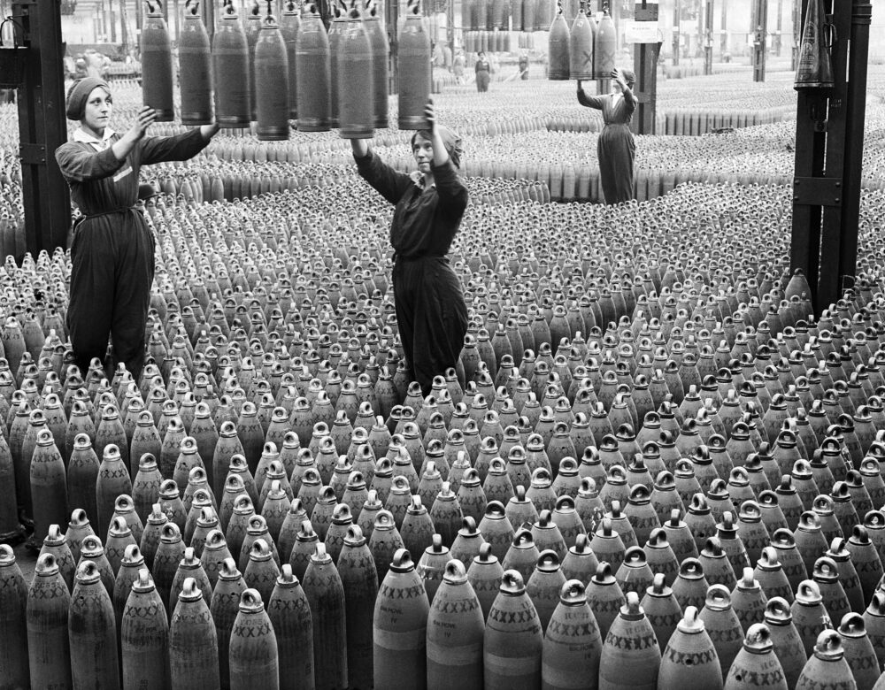 Old photo of women on a factory floor surrounded by munitions shells