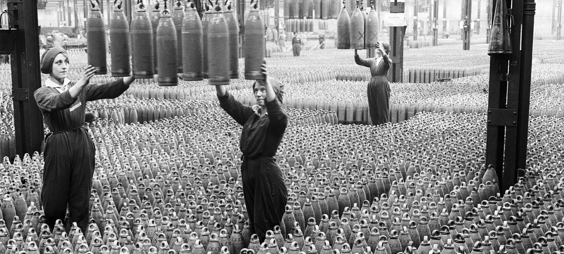 Old photo of women on a factory floor surrounded by munitions shells