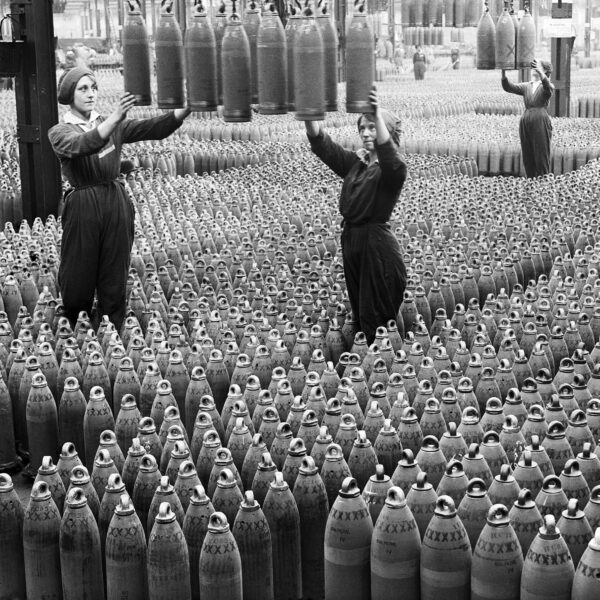 Old photo of women on a factory floor surrounded by munitions shells