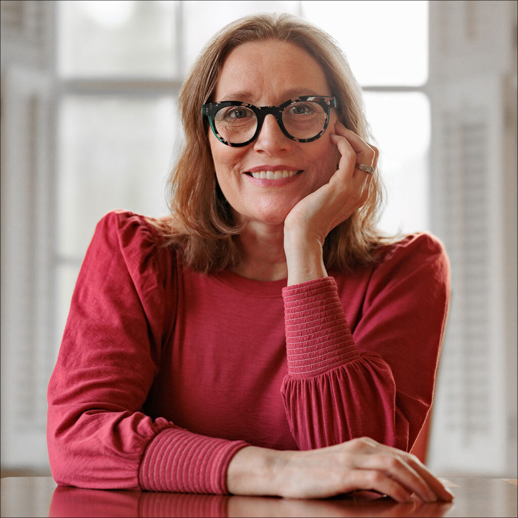 Beth Linker wearing glasses, dark pink top, chin on hand, seated at a table in front of a window