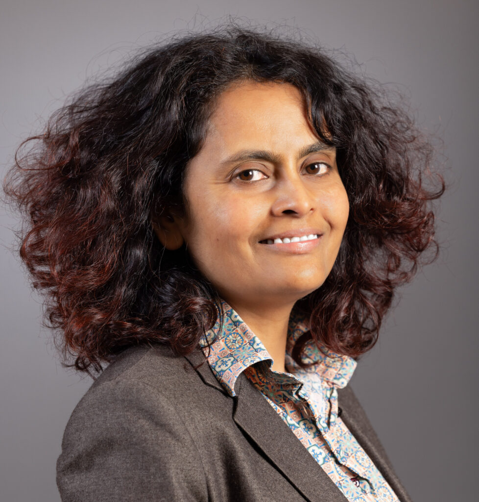 Surekha Davies, turned 3/4, smiling, in blazer and button-down shirt