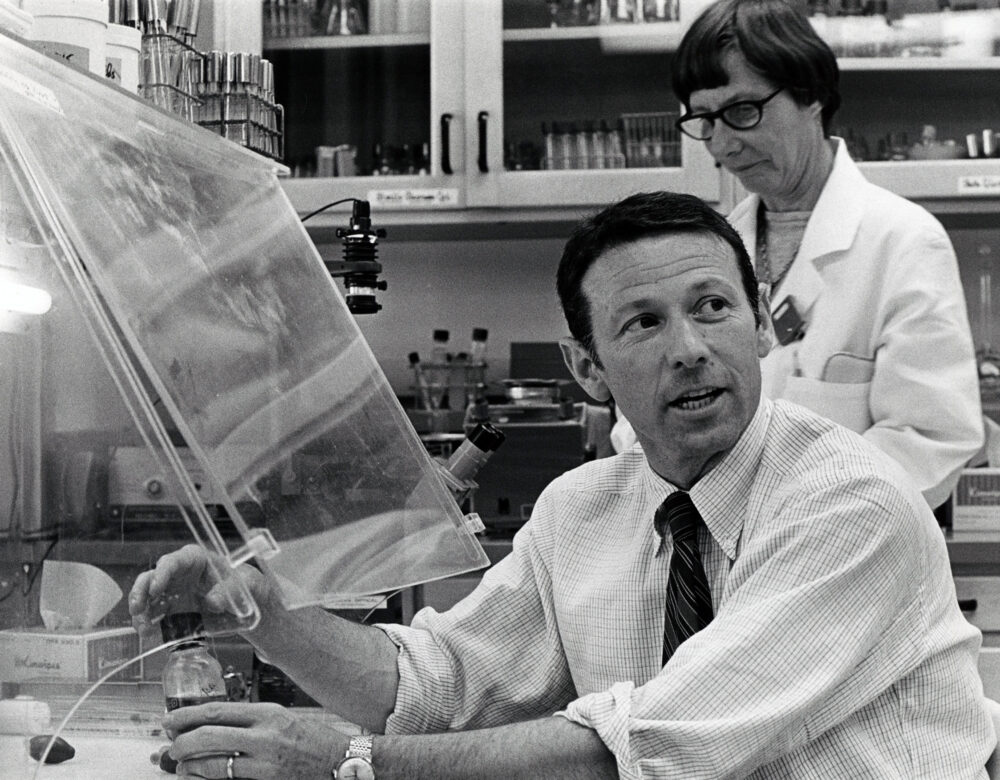 Man with rolled up shirt sleeves working under a lab hood while a woman in lab coat looks on