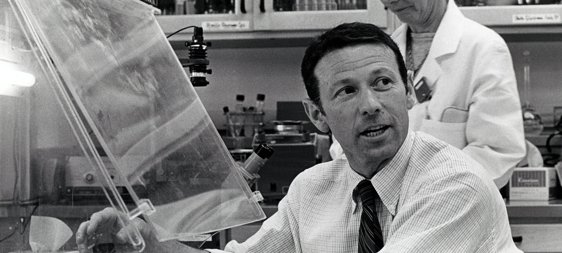 Man with rolled up shirt sleeves working under a lab hood while a woman in lab coat looks on