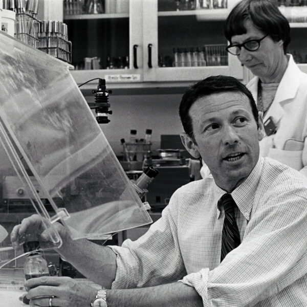 Man with rolled up shirt sleeves working under a lab hood while a woman in lab coat looks on