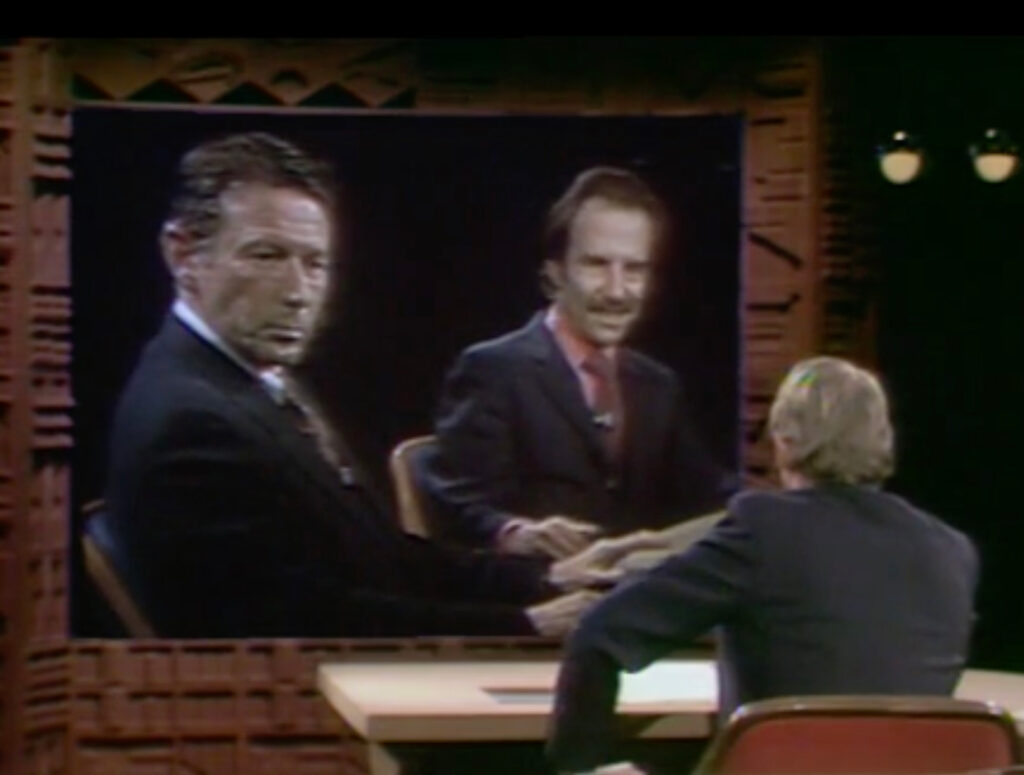 Interviewer talking to two men on a screen projector