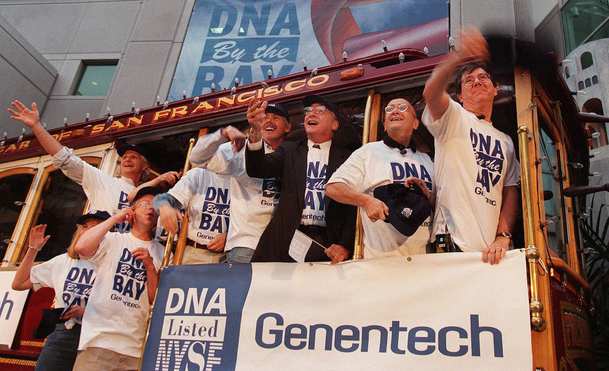 A crowd of people wearing t-shirts reading “DNA by the Bay” celebrate inside a trolley car