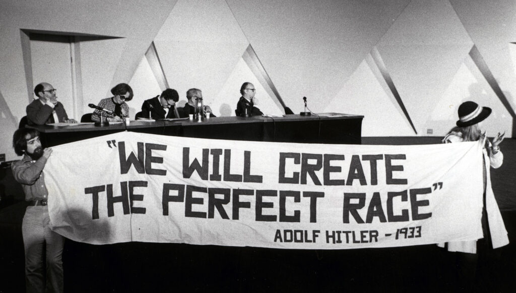 Two protesters hold a banner reading “we will create the perfect race, Adloph Hitler, 1933”